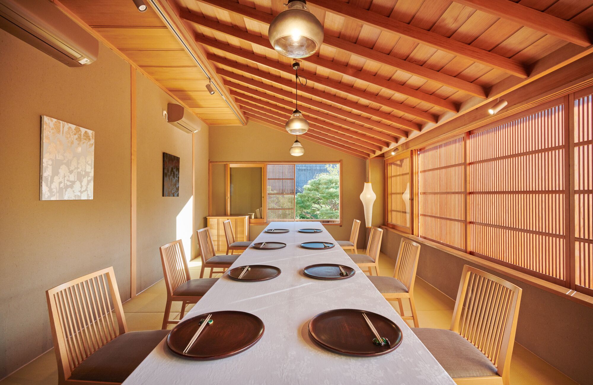 Uma sala de jantar tradicional japonesa com uma mesa comprida posta para quatro pessoas, vigas de madeira no teto e portas deslizantes à direita.