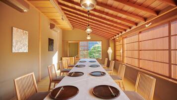 Uma sala de jantar tradicional japonesa com uma mesa comprida posta para quatro pessoas, vigas de madeira no teto e portas deslizantes à direita.