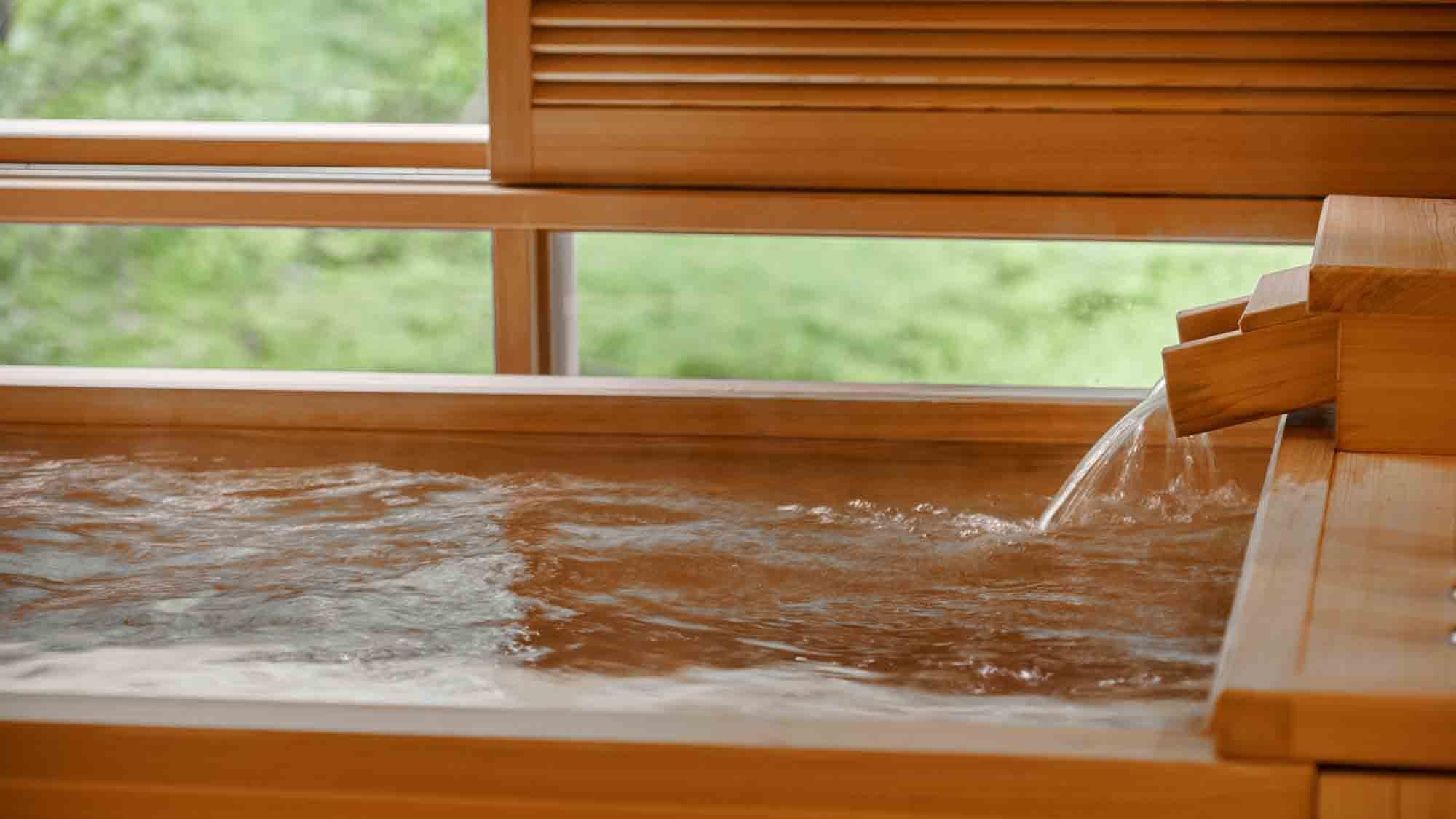 A wooden bathtub with a waterfall feature, set against a window with a view of greenery.