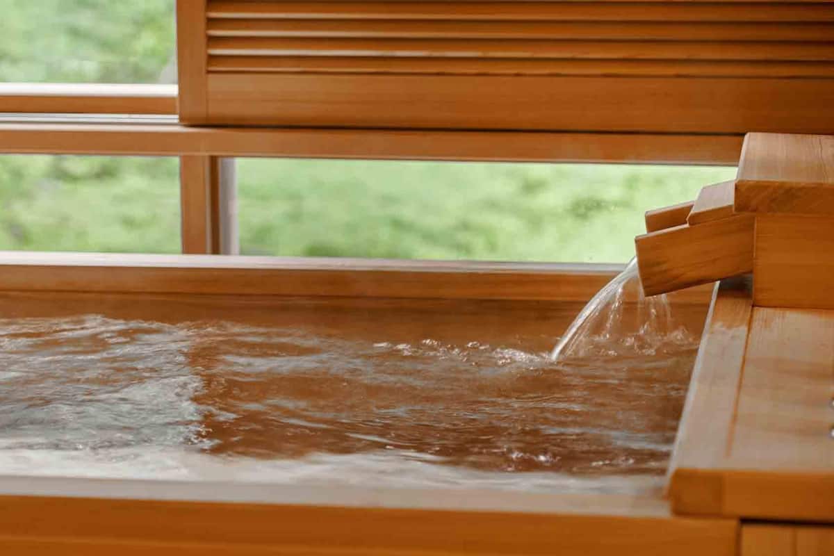 A wooden bathtub with a waterfall feature, set against a window with a view of greenery.