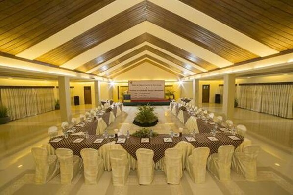 Un salón de banquetes con largas mesas dispuestas para un evento formal, con un arreglo floral central y un banderín en la parte delantera.