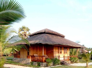 A wooden house with a thatched roof, surrounded by palm trees and a stone wall. - Sunny Paradise Resort (Ngwe Saung)