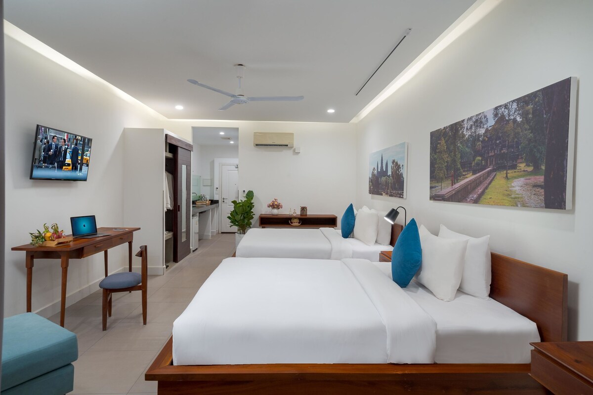 A modern hotel room with two beds, a desk with a laptop, a TV, and artwork on the walls.
