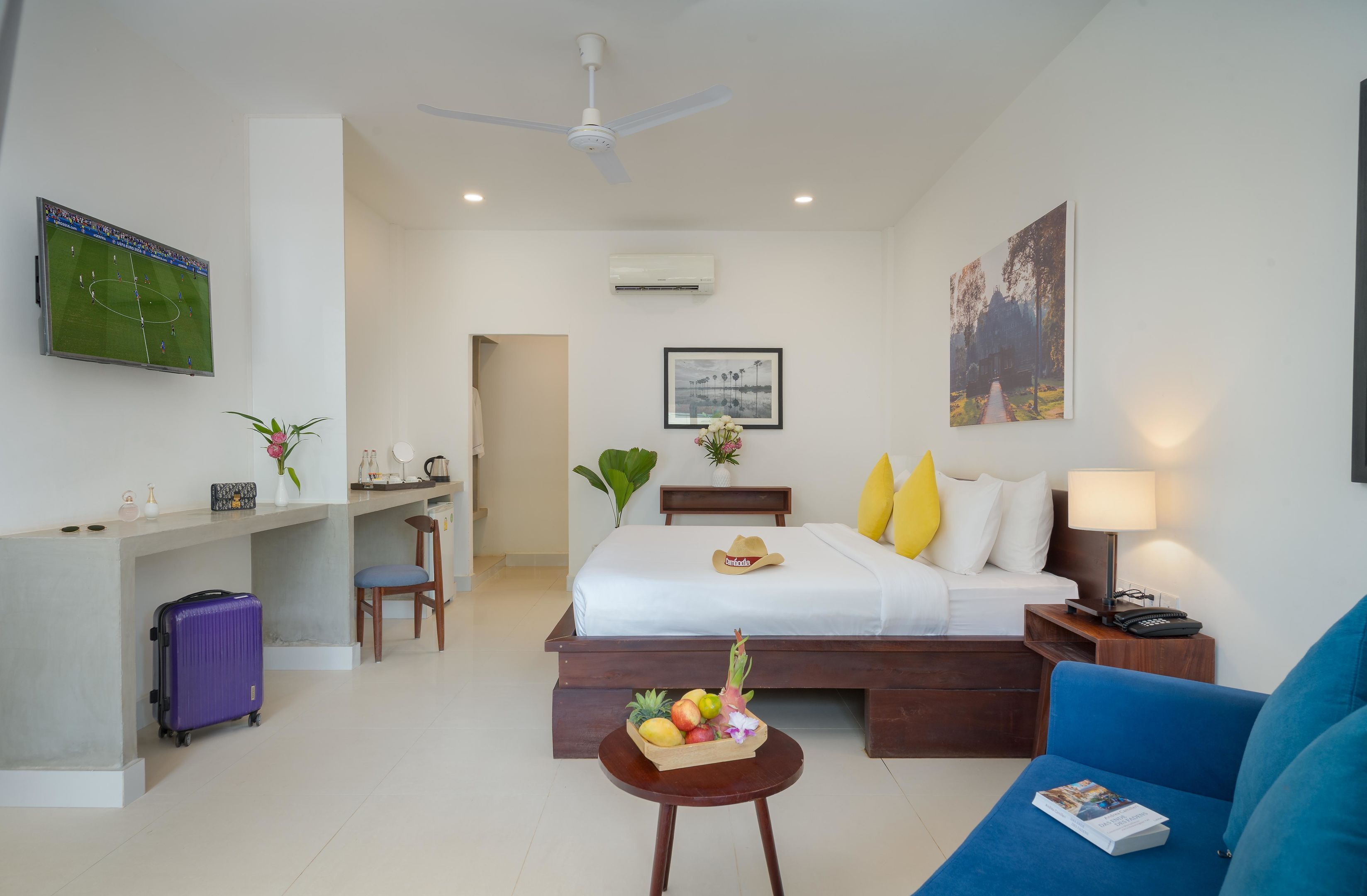 A hotel room with a bed, a desk, a chair, a sofa, a small table with fruit, and a ceiling fan.
