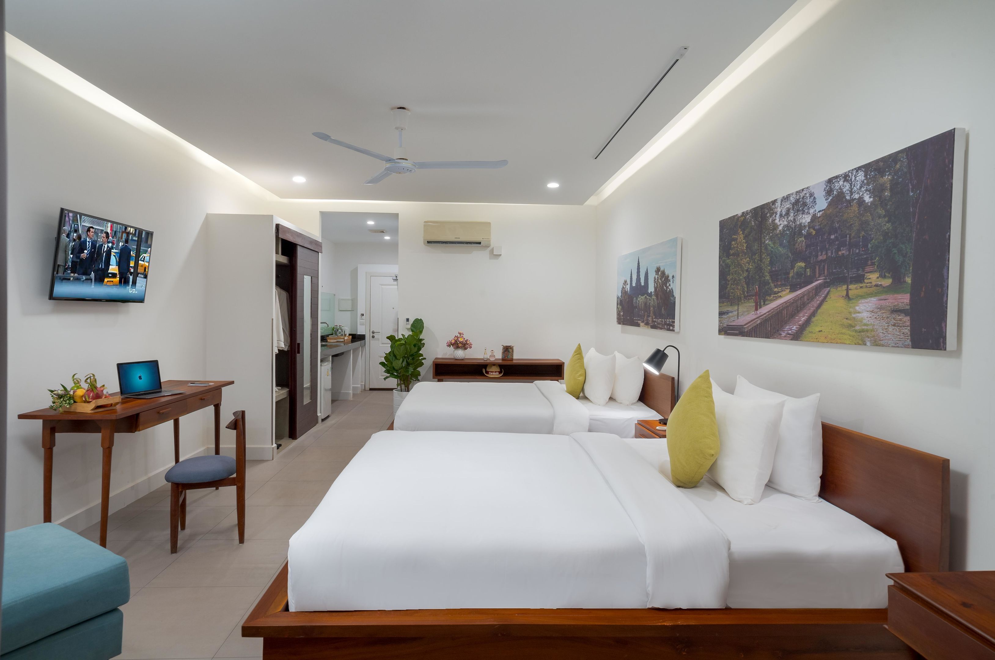 A hotel room with two beds, a desk with a laptop, a TV, and artwork on the walls.
