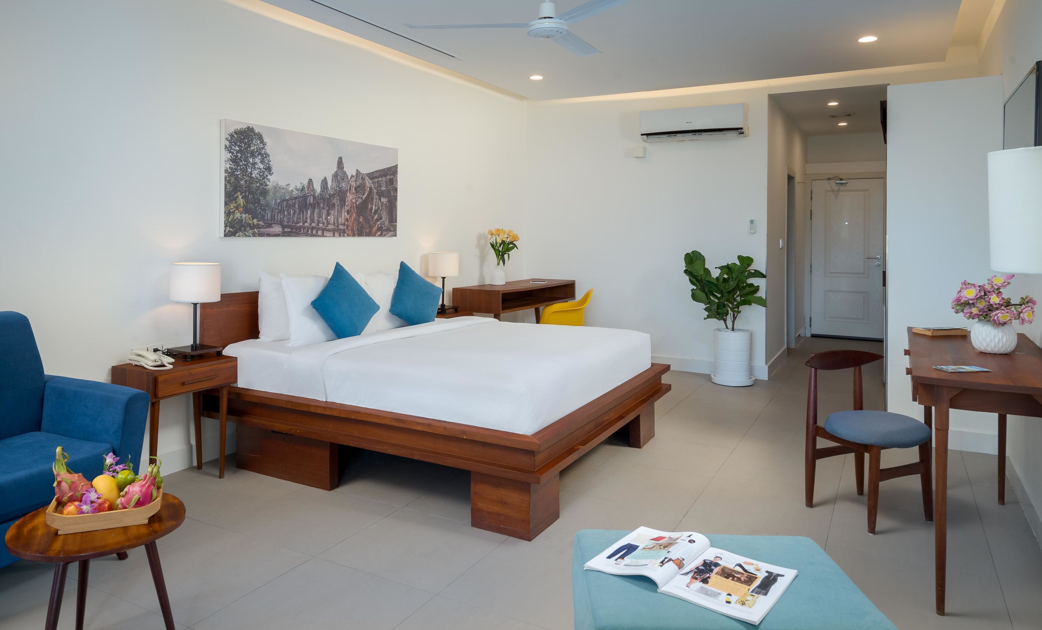 A modern hotel room with a wooden bed, a desk, a chair, a small table with a vase of flowers, and a wall-mounted air conditioner.