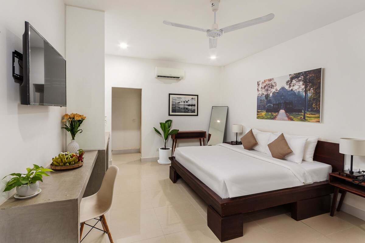 A modern hotel room with a bed, desk, chair, TV, ceiling fan, and artwork on the wall.