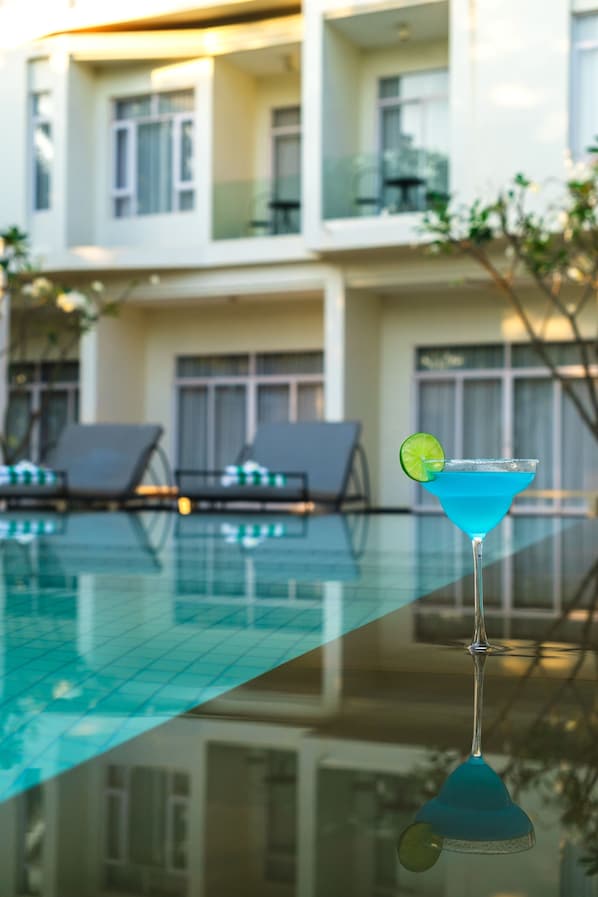 A clear glass pool with a blue drink on the edge, lounge chairs, and a building in the background.