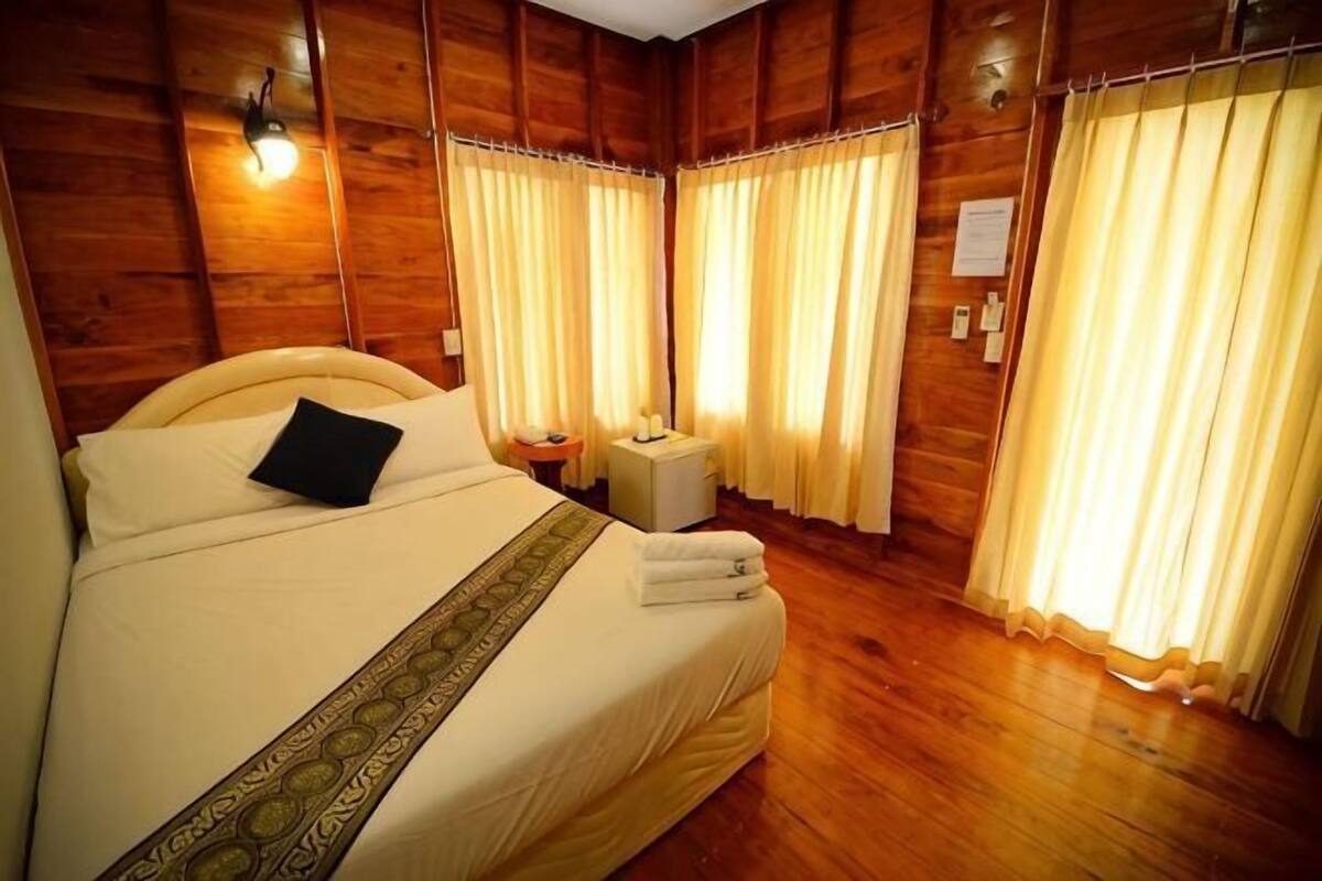 Una habitación de hotel con piso de madera, una cama con un almohadón, una mesita de noche y cortinas.