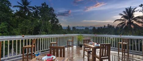 Un área de comedor al aire libre con mesas y sillas de madera, rodeada por una barandilla y con vistas a un paisaje tropical con palmeras y montañas.