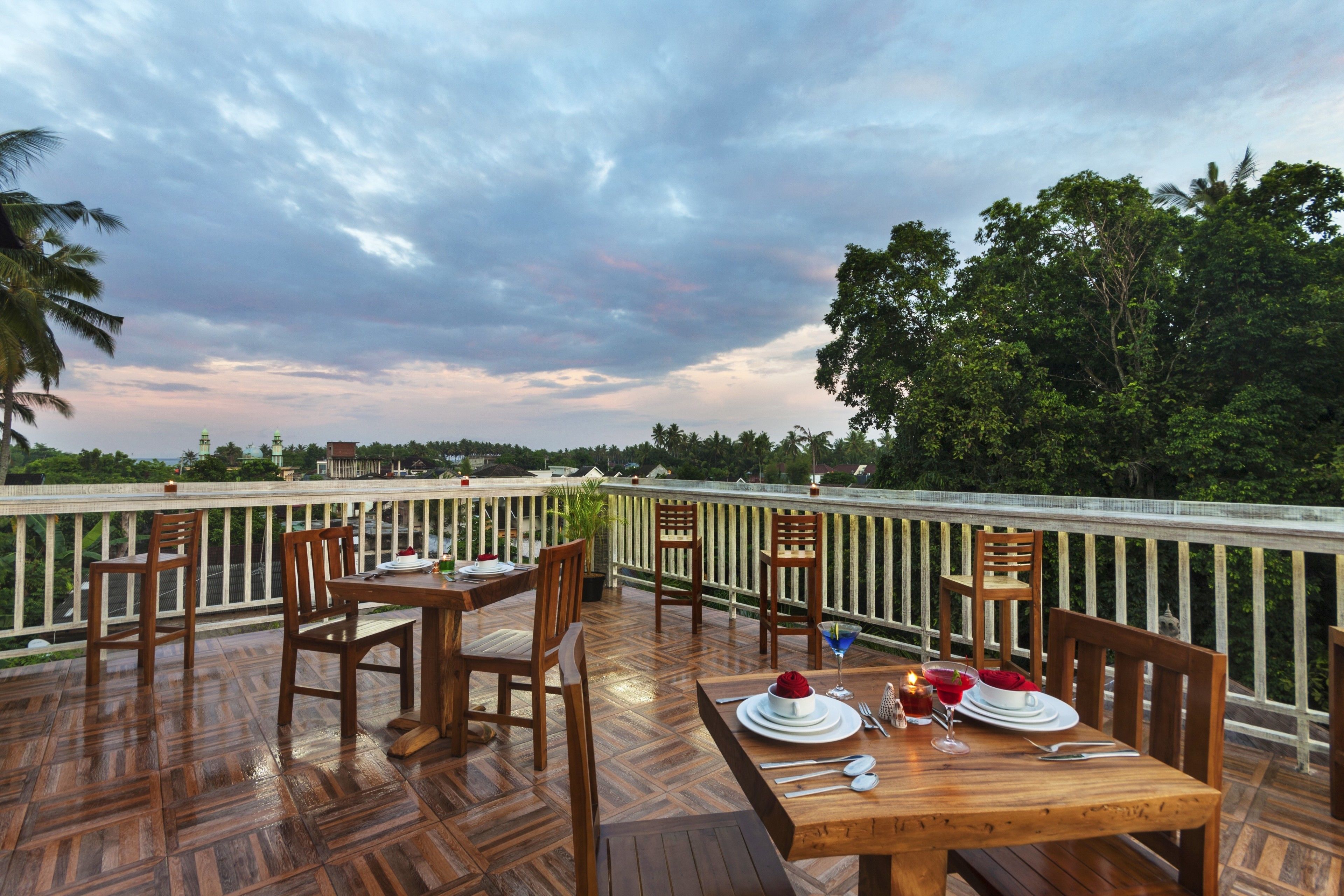 An outdoor dining area with wooden tables and chairs, set against a backdrop of a cloudy sky and lush greenery.
