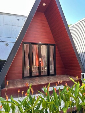 A triangular structure with a wooden floor, glass doors with curtains, and a small garden area with plants.