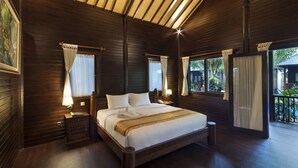 A large wooden-framed bed with white linens, two bedside tables with lamps, and a view of greenery outside.