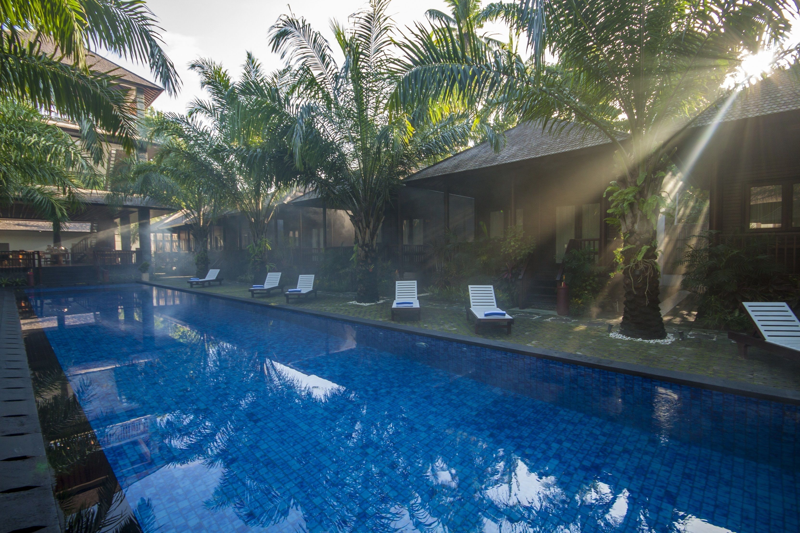 A swimming pool with lounge chairs, surrounded by palm trees and a building with a tiled roof.