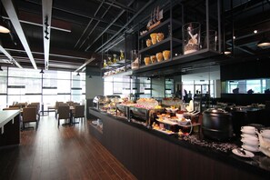 A buffet setup with various food items and beverages, a seating area with tables and chairs, and a modern interior design with exposed ceilings and large windows. - Nexus Regency Suites & Hotel (Shah Alam)