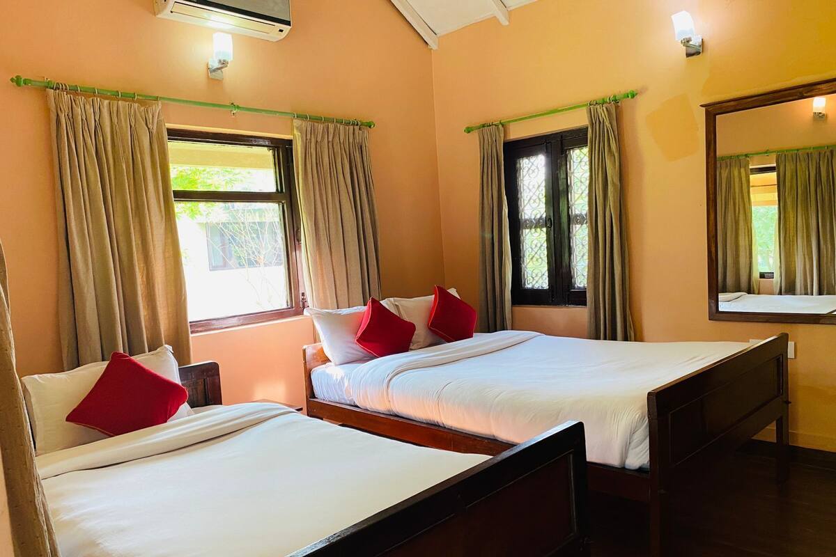 A room with two single beds, beige curtains, a window with a view, and a mirror on the wall.