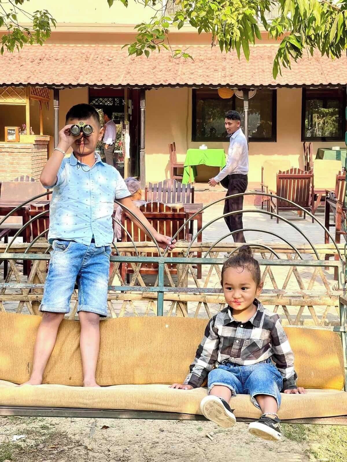 A child standing on a bench with a child sitting beside it, in front of a building with a tiled roof and outdoor seating.