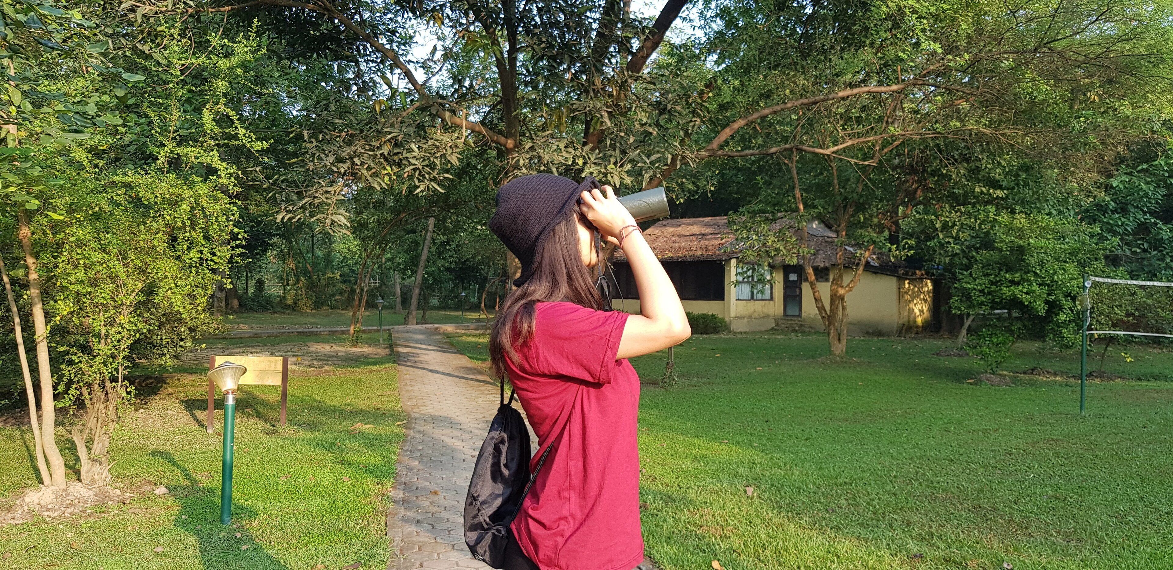 A person in a red shirt and black hat walking along a paved path in a park with trees and a small building in the background.