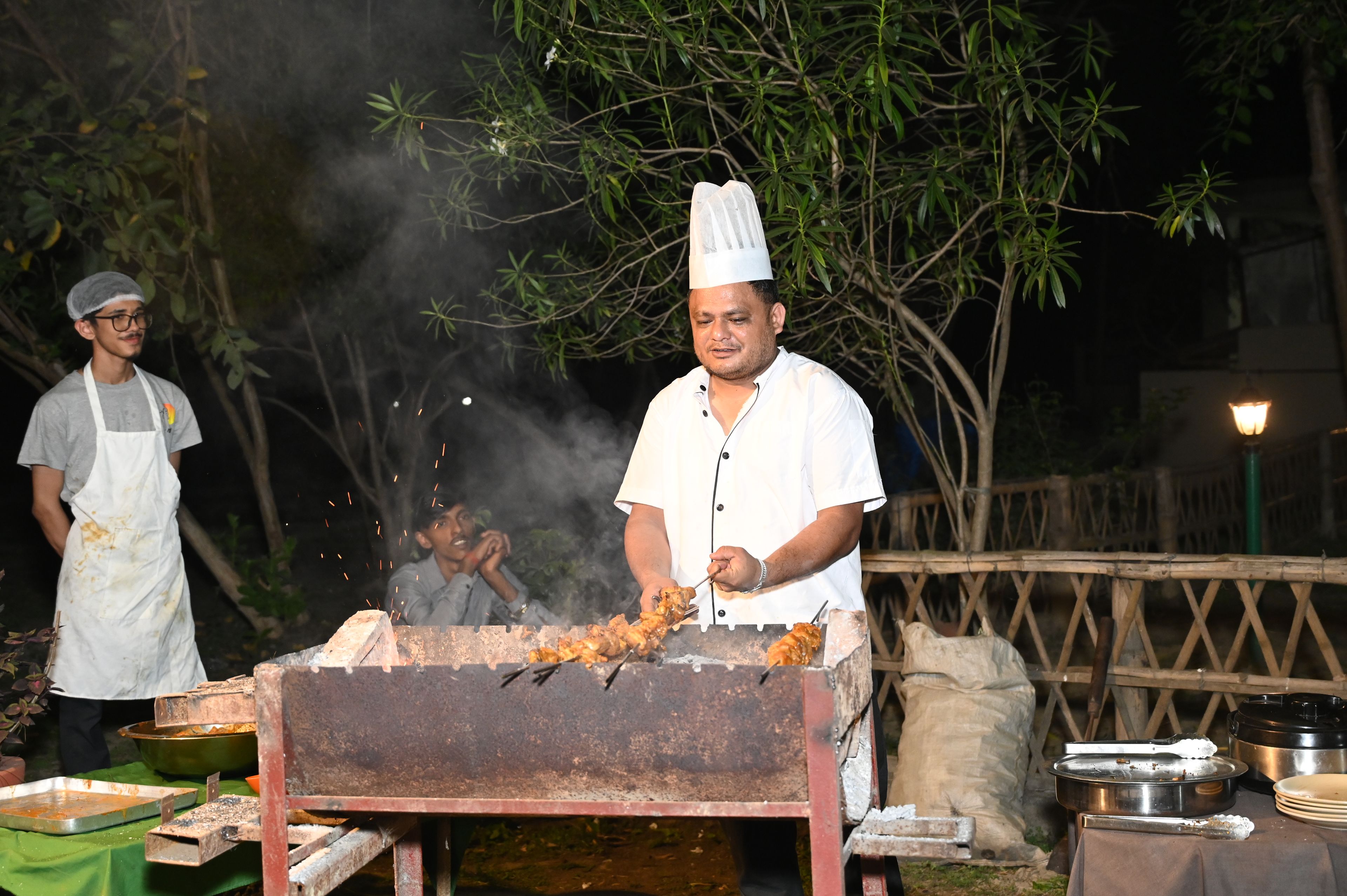 Un chef asando comida al aire libre, con una parrilla y utensilios de cocina a la vista.