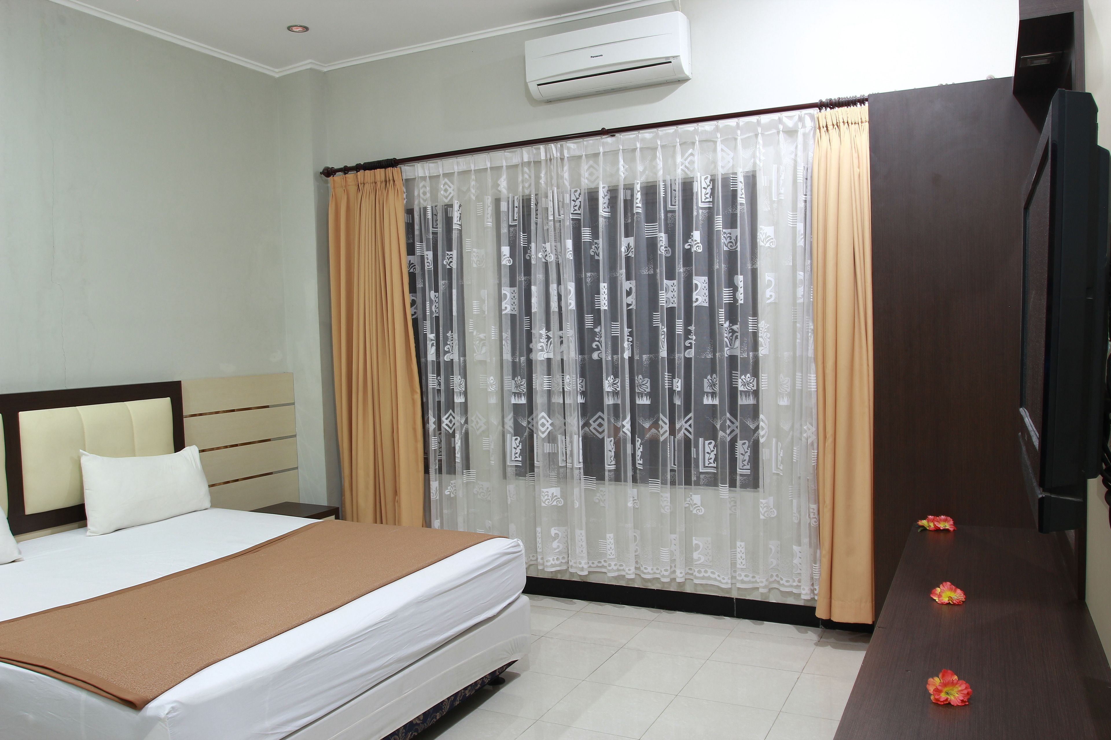 A hotel room with a bed, a wooden nightstand, a TV, and a window with patterned curtains.
