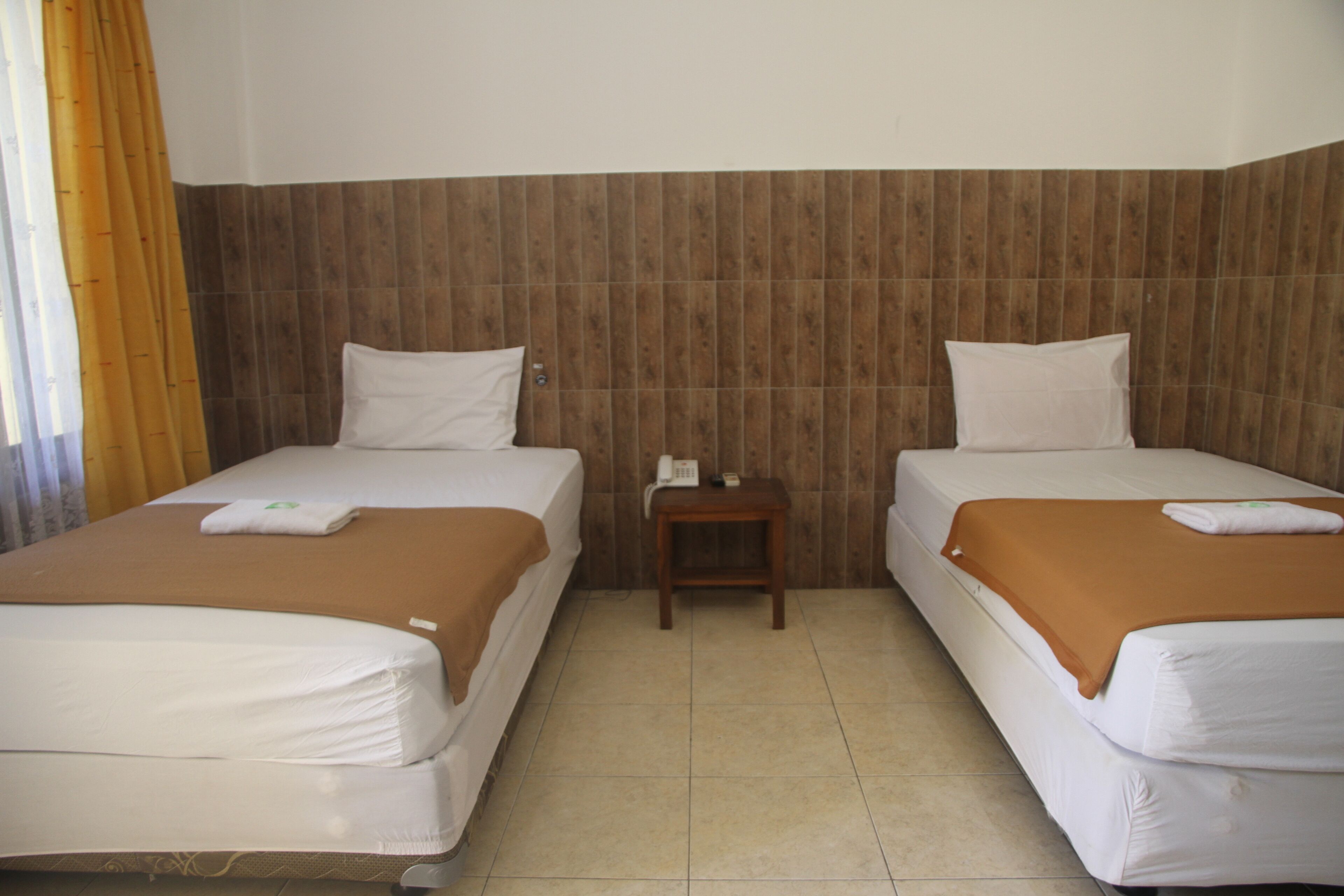 A hotel room with two beds, a wooden headboard, a small bedside table, and a curtain.