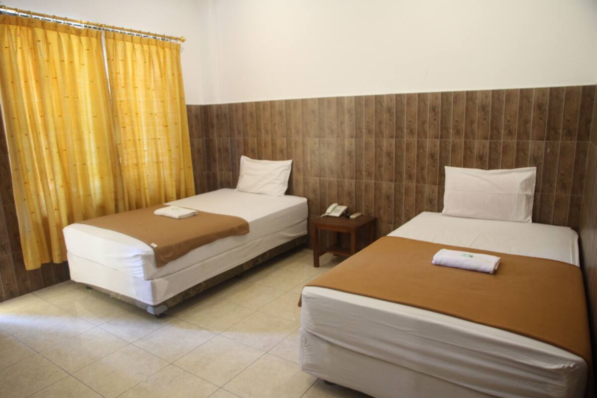 Une chambre d’hôtel avec deux lits, une tête de lit en bois, une petite table de chevet avec un téléphone et un rideau.
