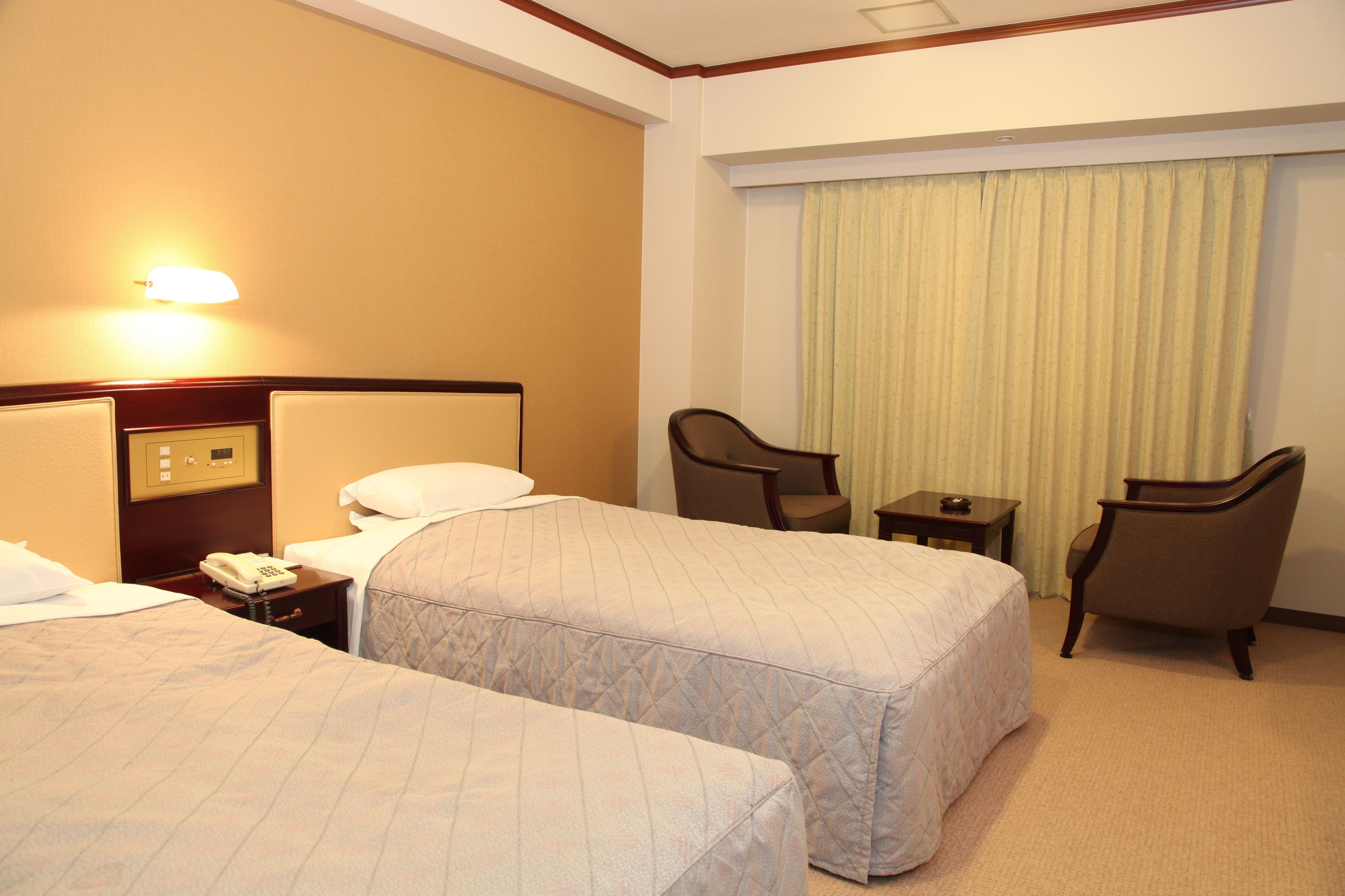 A hotel room with two beds, a desk with a telephone, two chairs, and a window with curtains.