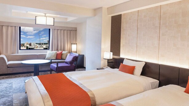 Een hotelkamer met twee bedden, een bank, een bureau en een groot raam met uitzicht op de stad.