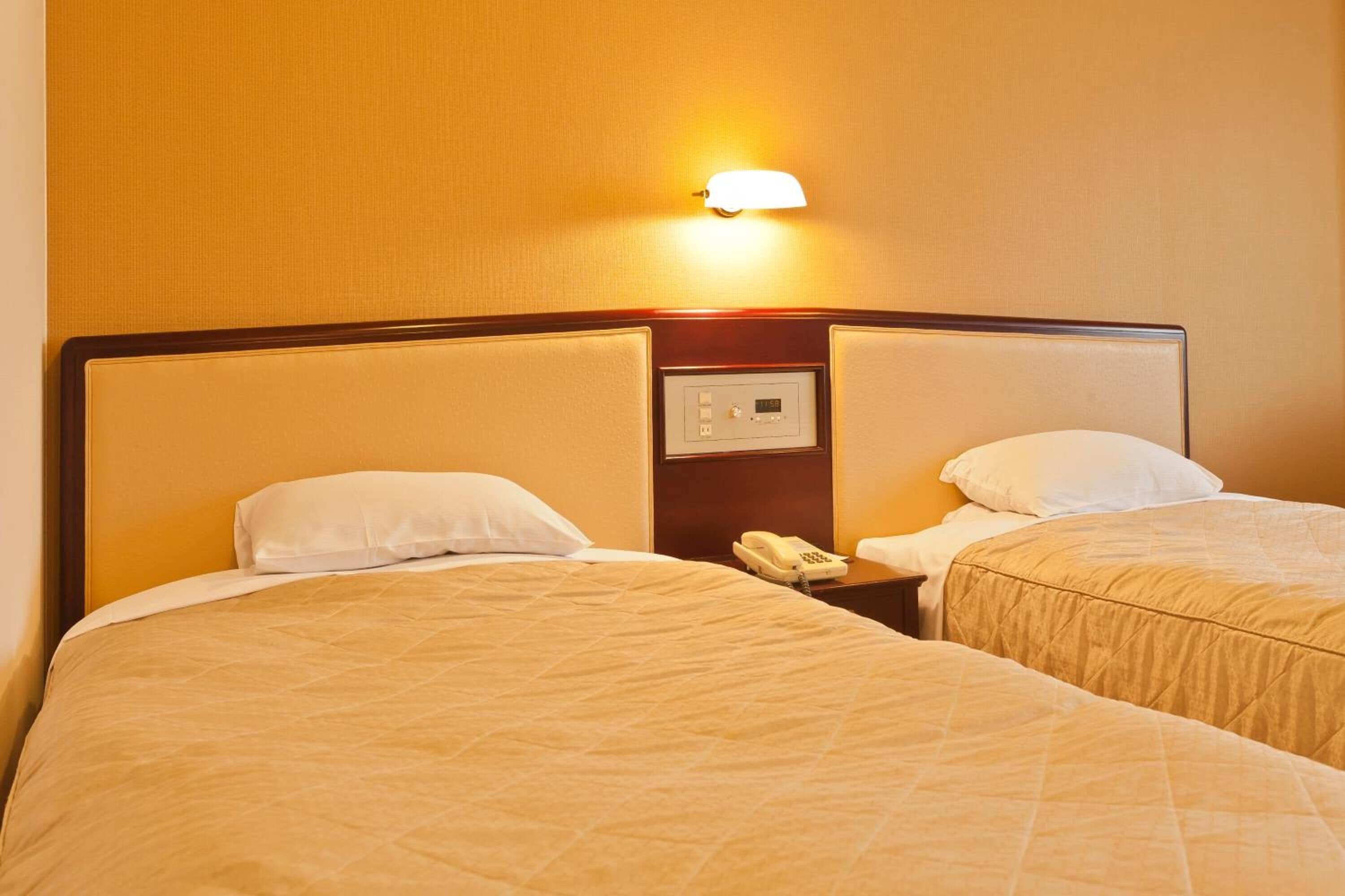 A hotel room with two beds, each with a headboard, a bedside table with a phone, and a wall-mounted light fixture.