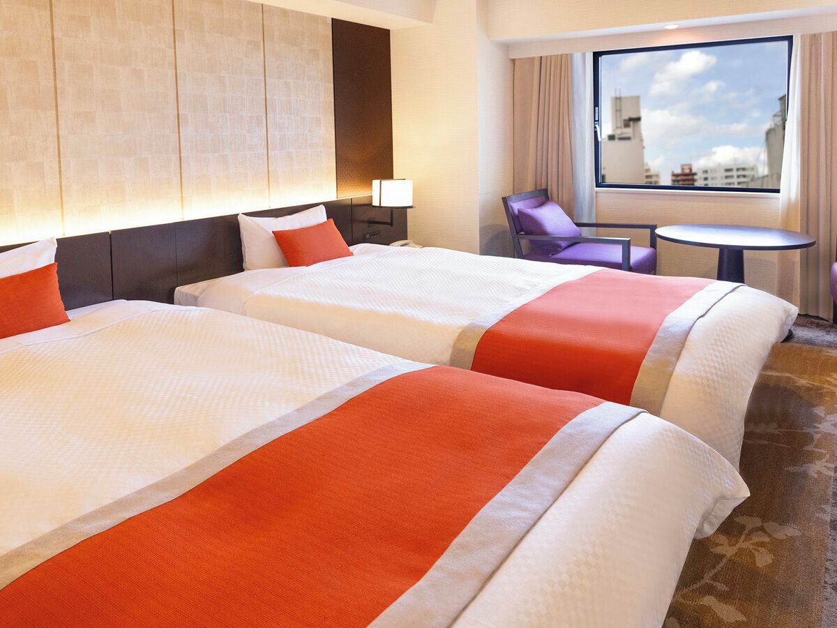 A hotel room with two beds, a small table, a chair, and a window with a city view.