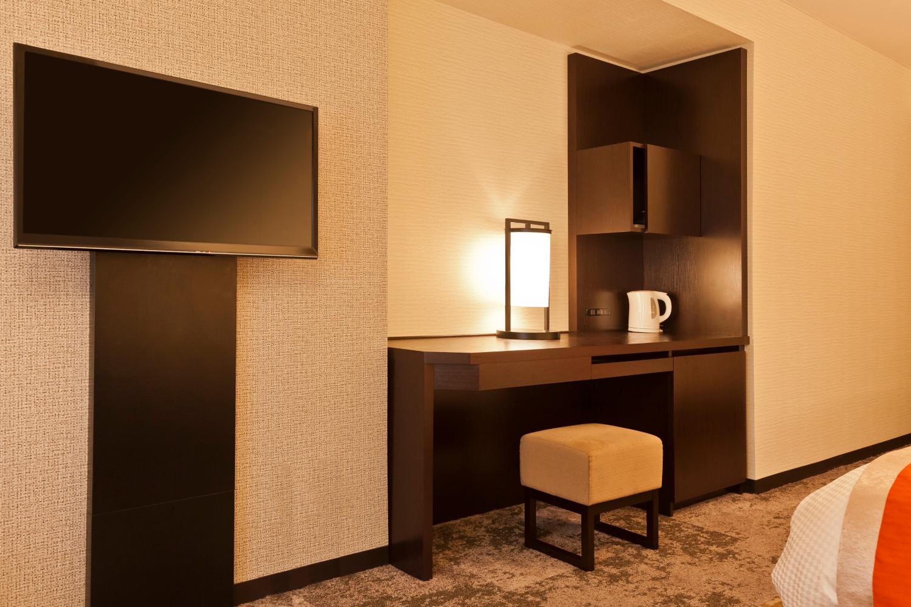 A hotel room with a flat-screen TV, a desk with a lamp, a coffee maker, and a small stool.