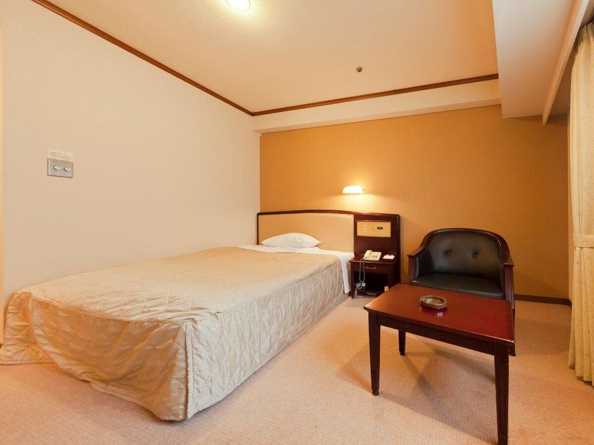 A hotel room with a single bed, a bedside table with a lamp, a chair, and a small table with a tray.