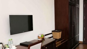 Quarto com televisão de tela plana na parede, escrivaninha de madeira com cesto e guarda-roupa com portas venezianas.