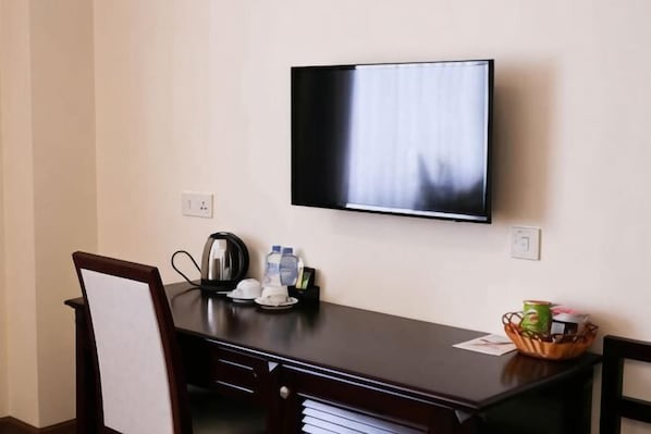 A hotel room with a flat-screen TV mounted on the wall, a wooden desk with a kettle, cups, and a basket of snacks.