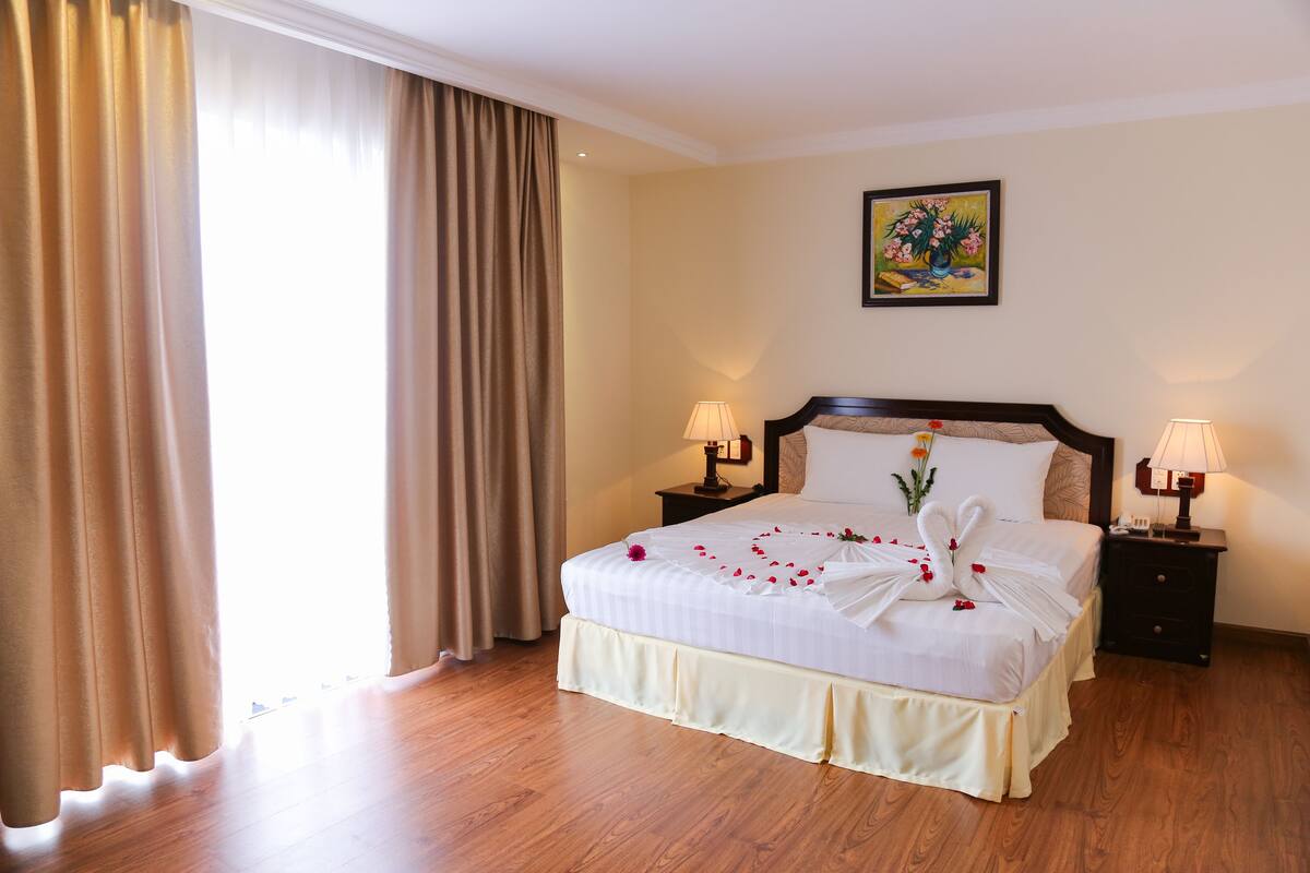 A hotel room with a large bed, two bedside tables with lamps, a painting on the wall, and a window with curtains.
