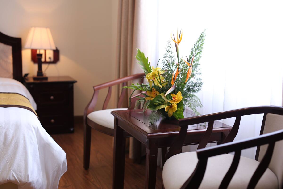 A hotel room with a bed, a nightstand with a lamp, a wooden chair, and a table with a floral arrangement.