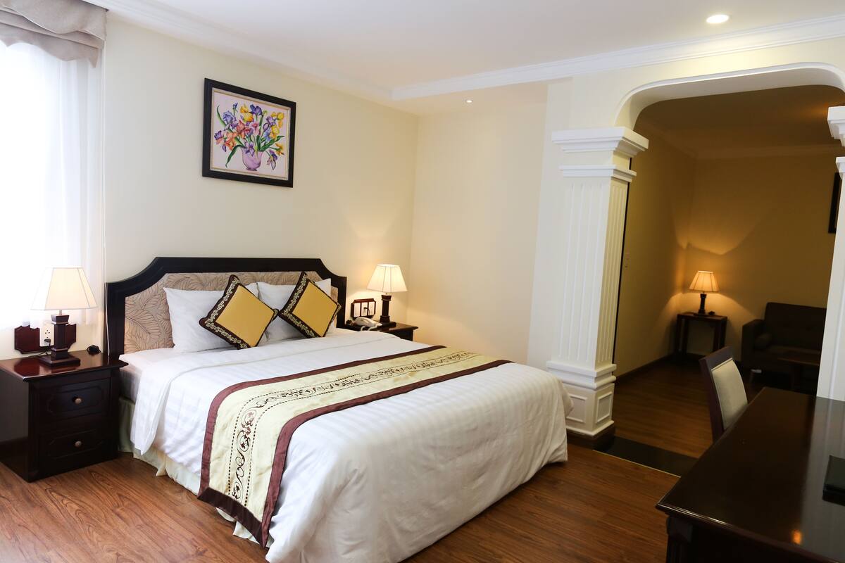 A hotel room with a large bed, two bedside tables with lamps, a framed floral artwork on the wall, and a wooden floor.