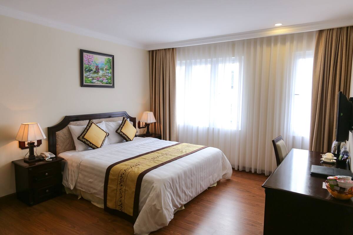 A hotel room with a large bed, two bedside lamps, a nightstand, a desk with a chair, and a window with curtains.