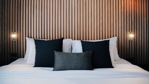 Una cama bien hecha con sábanas blancas y dos almohadas, frente a una pared de listones de madera con dos lámparas fijadas a la pared.