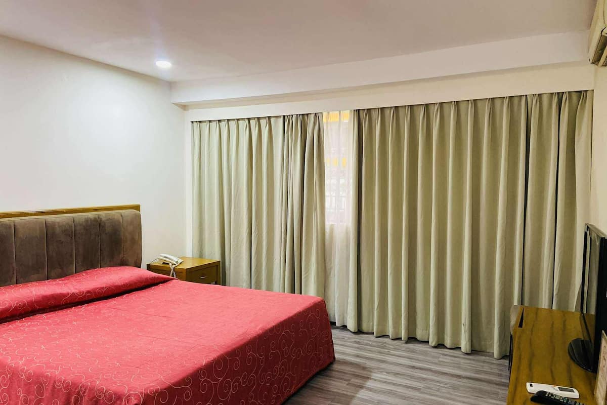 A hotel room with a large bed, a bedside table with a phone, a wooden desk with a TV and remote, and a window with curtains.
