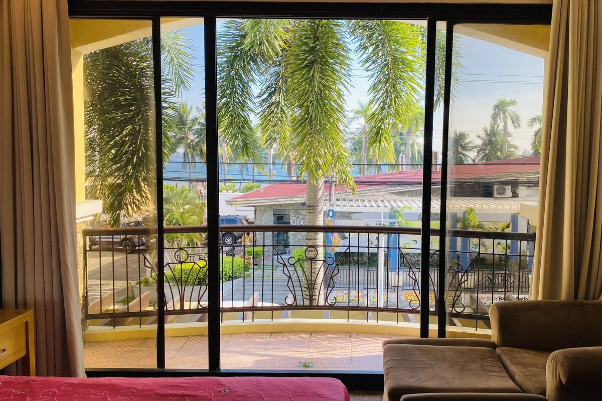 A room with a large window, a balcony with a metal railing, a brown sofa, and a small table.