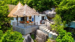 Una casa con techo de paja y terraza de madera, rodeada de exuberante vegetación y grandes rocas.