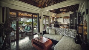 Amplia sala de estar con un sofá grande, una mesa de centro de madera y una escultura. Hay una puerta de cristal que da a un área de estar exterior con vistas al paisaje.