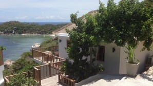 Una propiedad costera con terraza de madera, un edificio pequeño con balcón y vista al mar y colinas.