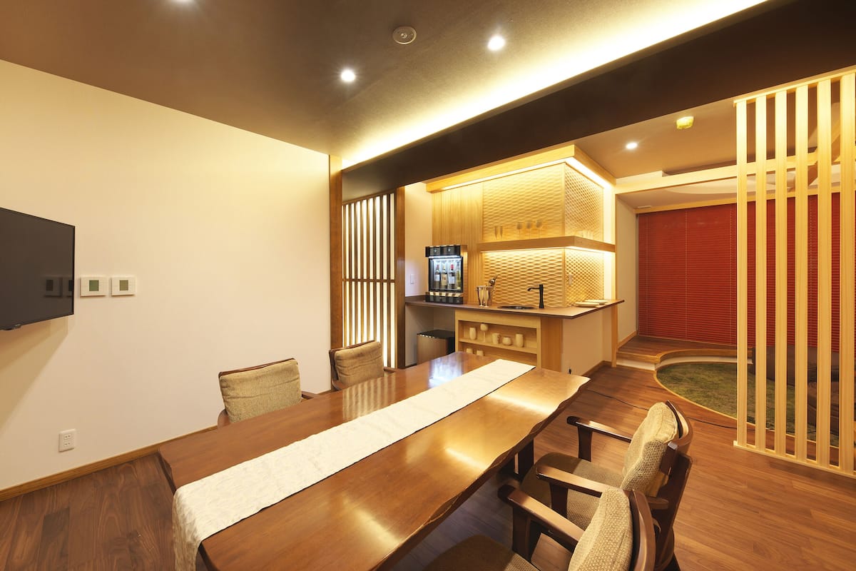 A modern dining area with a long wooden table, chairs, and a kitchen area with a refrigerator and counter.