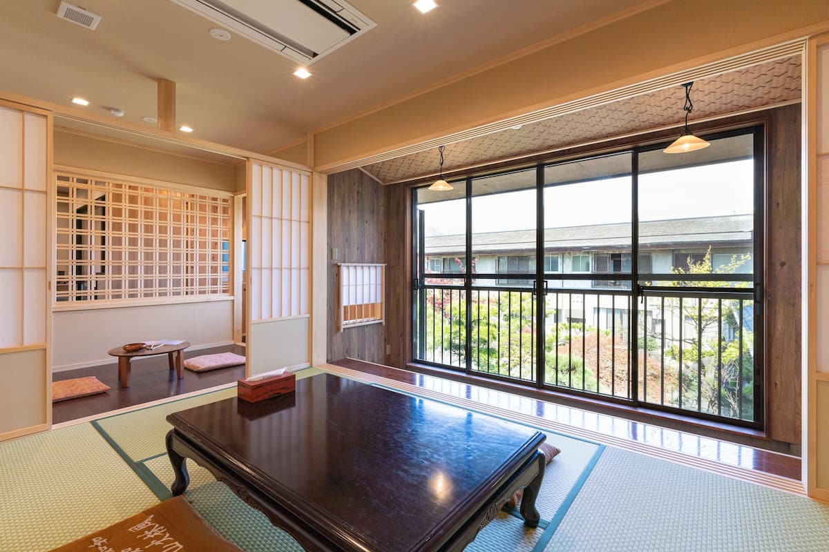 A traditional Japanese-style room with tatami flooring, a low wooden table, sliding doors, and a large window offering a view of a residential area.