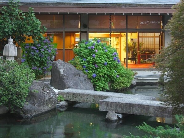 Un jardín japonés tradicional con un puente de piedra sobre un estanque, abundante vegetación y un edificio con grandes ventanales al fondo.