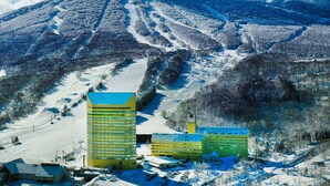Una montaña nevada con pistas de esquí y un telesilla. Se observan varios edificios con techos planos y un estacionamiento con coches.