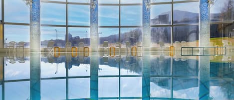An indoor swimming pool with clear glass walls, multiple chairs, and a view of the outdoors.
