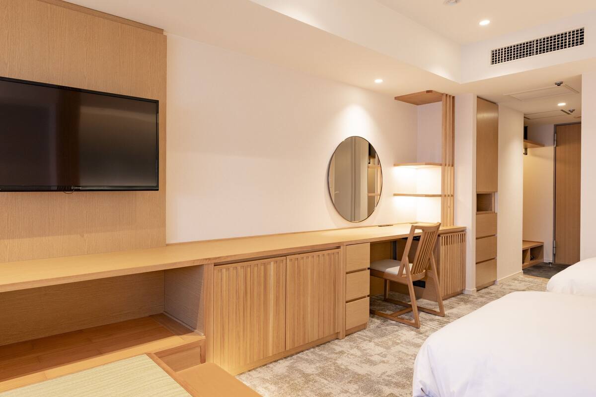 A hotel room with a flat-screen TV mounted on the wall, a desk with a chair, a round mirror, and a bed.